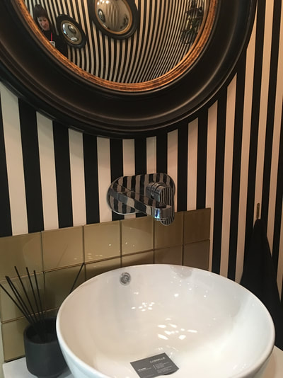 Lavatory with black and gold, convex wall mirror on black and white, vertical striped wallpaper with gold glass tiles