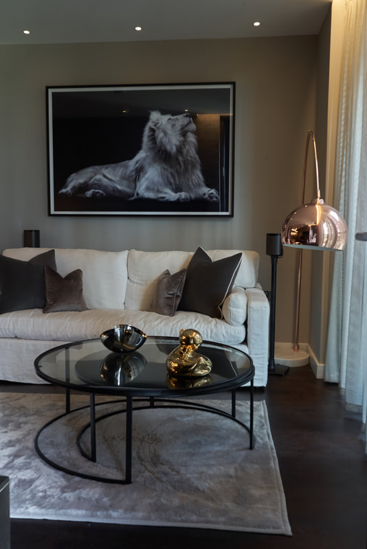 Living room with floor-standing, copper lamp, taupe sheers o the window, lion artwork, cream, linen sofa and glass coffee tables