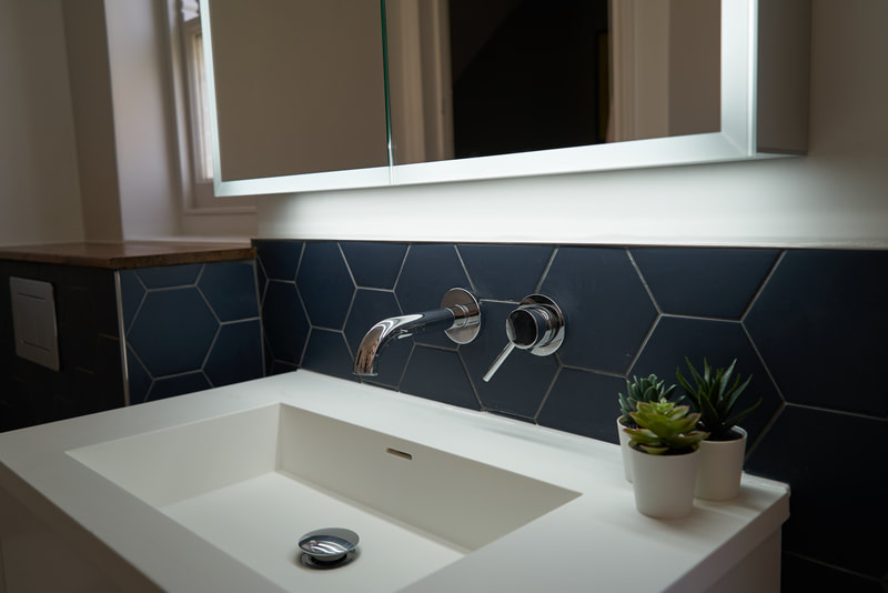 White, minimal, composite stone sink from Lusso Stone with anavy hexagonal tile splashabck from Claybrook and an LED bathroom cabinet from Drench