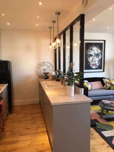 Open plan pastel colours living room and grey, painted kitchen separated by half glazed divider with a large portrait of Cara Delavigne